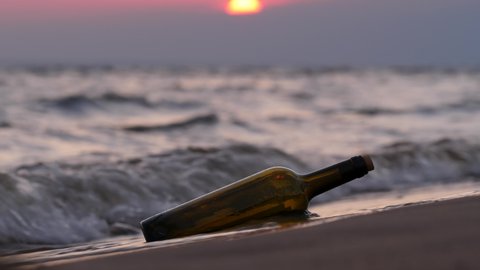 Bottle with message thrown ashore lies and then swayed from splash of oncoming wave. Close up shot of glass bottle traveled across sea to unknown beach, blurred sunset visible on background