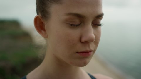 Closed eyed calm girl meditating on beach close up. Portrait of attractive girl breathing deeply outdoors. Concentrated lady face in front grey sea. Pretty sportswoman exhaling practicing yoga.