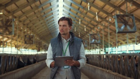Livestock owner checking animal feedlots making notes on clipboard at cowshed. Handsome man focused agricultural worker walking barn inspecting dairy facility. Milking manufacture professional concept