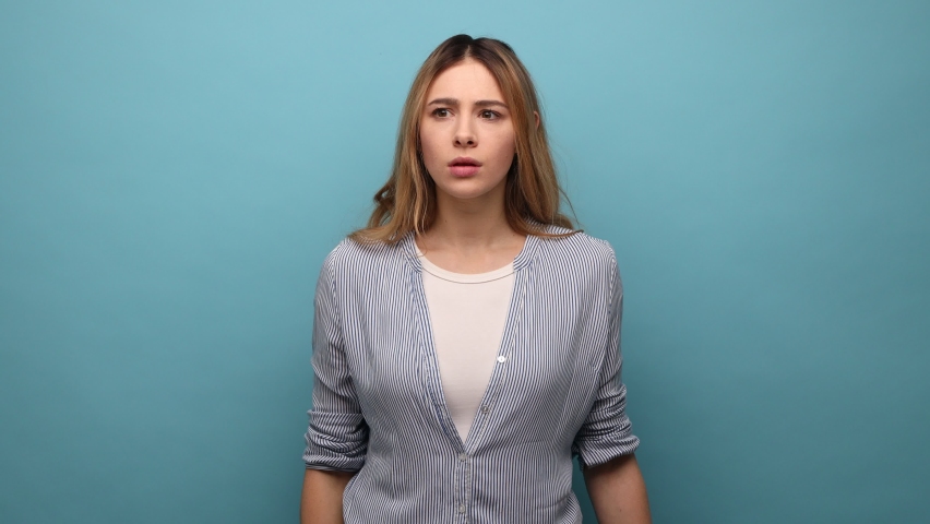 Curious woman holding hand above eyes and peering into distance, looking far away, expecting and searching someone on horizon, wearing striped shirt. Indoor studio shot isolated on blue background. Royalty-Free Stock Footage #1089988309