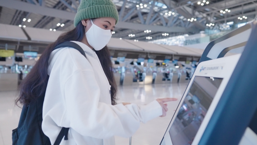 Asian female traveler wear face mask using airport automatic kiosk machine to check in for flight ticket, new technology for public transportation, online digital resources, self touch screen machine