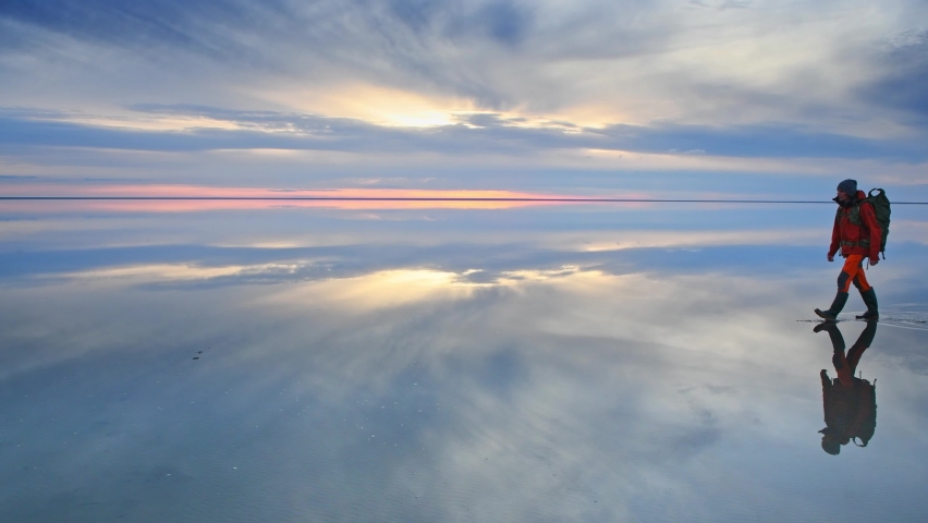 Man traveler with backpack walking on the salt lake at sunset. Blue sky with clouds are reflected in the mirror water surface. Travel and adventure concept
