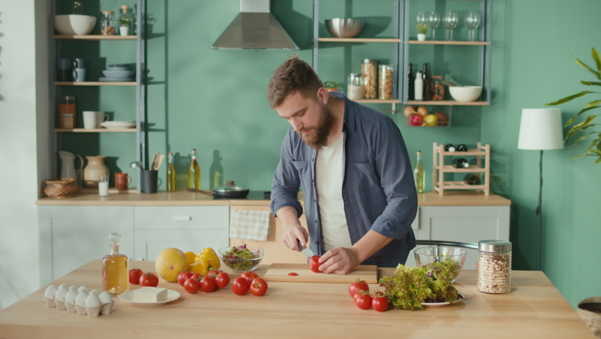 Cheerful Man With Beard Cutting Tomato on Cutting Board in His Kitchen. Handsome Young Man Cooking Vegetarian Breakfast Cutting Vegetables for Salad in Kitchen at Home. Healthy Lifestyle. Royalty-Free Stock Footage #1089993307