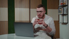 The guy in glasses holding a cup in his hands responds to information on a laptop