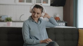Young happy woman is sitting on the chair, listening to music with headphones and singing. Home entertainment concept. Slow motion video.
