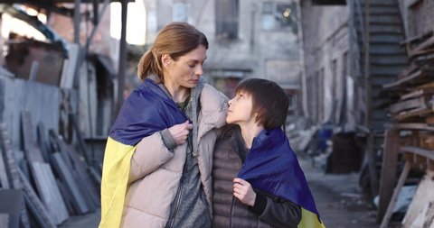 Upset little boy teen standing outdoor with sad tired mother covered in Ukrainian flag looking at camera while standing on street in ruined destroyed city. Family, refugees, patriot, patriotic concept