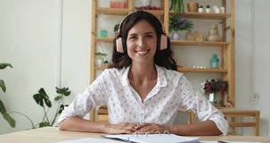 Young Hispanic woman wear wireless earphones sit at desk look at camera smile wave hand hi chat to friend by video call hold motivation speech take part at web conference. Screen web cam view portrait