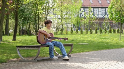 Lonely teenager boy sitting on a bench play guitar boring and entertaining himself