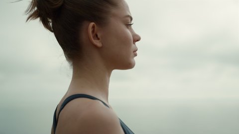 Profile of sporty girl turning face to camera standing outdoors cloudy day close up. Serious woman practicing yoga breathing exercise outside. Confident calm model posing on sea beach summer morning. 
