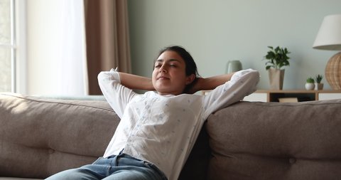 Peaceful relaxed young single Indian female enjoy healthy tranquil rest sitting on comfy sofa at home putting hands over head. Calm millennial woman recline on couch close eyes breath cool fresh air