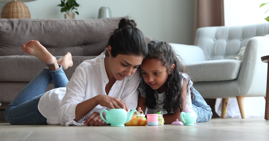 Happy young Indian mother female babysitter lying on warm heated floor at living room pretend eating toy pastries with little girl. Mommy and child daughter playing tea party game using plastic teaset Royalty-Free Stock Footage #1090008973