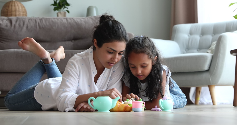 Happy young Indian mother female babysitter lying on warm heated floor at living room pretend eating toy pastries with little girl. Mommy and child daughter playing tea party game using plastic teaset Royalty-Free Stock Footage #1090008973