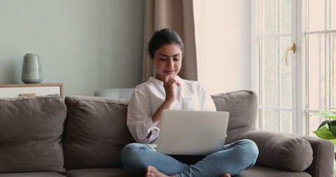 Smiling young Indian woman sit cross legged on cozy sofa hold notebook computer on laps chat in messenger think before write answer. Happy female student relax at home use entertainment app on laptop