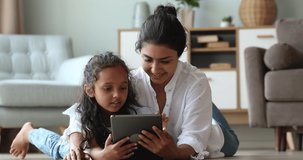 Friendly Indian foster mother little adopted daughter lie rest on warm floor at living room use online app on electronic pad. Smiling adult person nanny elder sister play web game with child on tablet