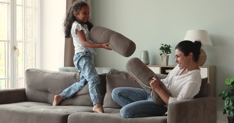 Grownup elder sister play with younger girl child on cozy couch imitate funny battle act shooting using sofa cushions as guns. Laughing Indian mom enjoy imaginary fight on pillows with little daughter
