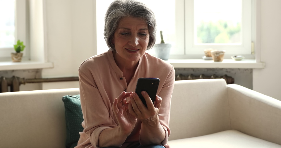Happy smiling middle aged Hispanic woman on pension rest on couch with smartphone scroll web pages swipe photos on dating website surf news at social media. Beautiful aged lady using phone application Royalty-Free Stock Footage #1090009079