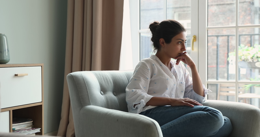 Thoughtful sad Indian female sit in armchair by window at living room ponder on important choice try to make correct decision in difficult life situation. Anxious young lady feel uncertain hesitate Royalty-Free Stock Footage #1090009095