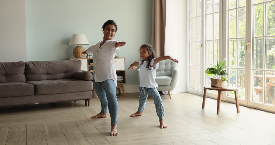 Happy friendly Indian family young mum little daughter schoolgirl hold warrior two yoga pose at light cozy living room at morning. Excited small girl imitate sporty mommy yogi learn to do easy asana Royalty-Free Stock Footage #1090009101