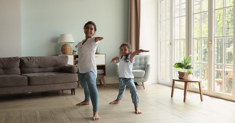 Happy friendly Indian family young mum little daughter schoolgirl hold warrior two yoga pose at light cozy living room at morning. Excited small girl imitate sporty mommy yogi learn to do easy asana