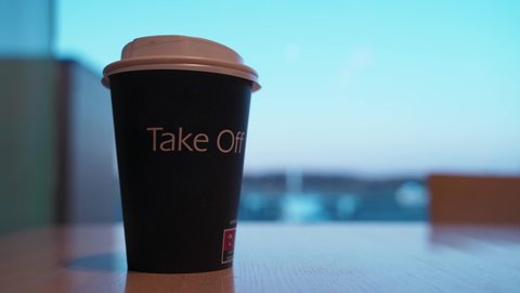 Man's hand putting a paper cup of coffee on a table at the airport. Airplane on the background behind a big window. Focus is pulling from airplane to the cup