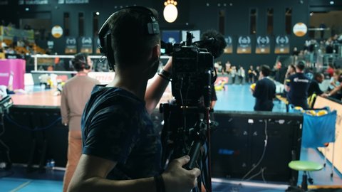 Camera Operator On TV Broadcasting Production Channel. Camera Operator On Sports Shoot Media Movie. Cameraman Working In Volleyball Area. Making TV Sport Show. Women Volleyball Team on Blurred