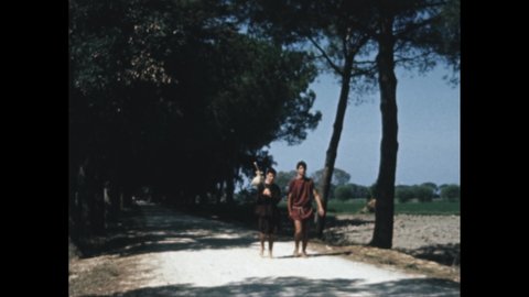 Decade 1950s: young Roman man in white toga. Roman age people walk along road.