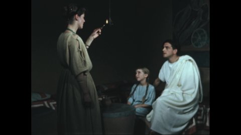 1950s 100 BC: Woman wears robe, lights hanging lamp, blows out match. Man, girl, sit, watch. Boy wears toga, sandals, stands proudly, smiles.