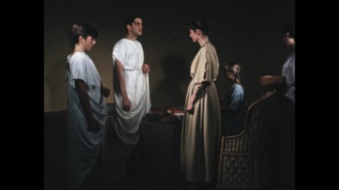 1950s 100 BC: Man stands, wears toga, talks. Woman turns, gives bread to person, pats girl on back. Boy listens, nods. Man leans over counter, talks, smiles.
