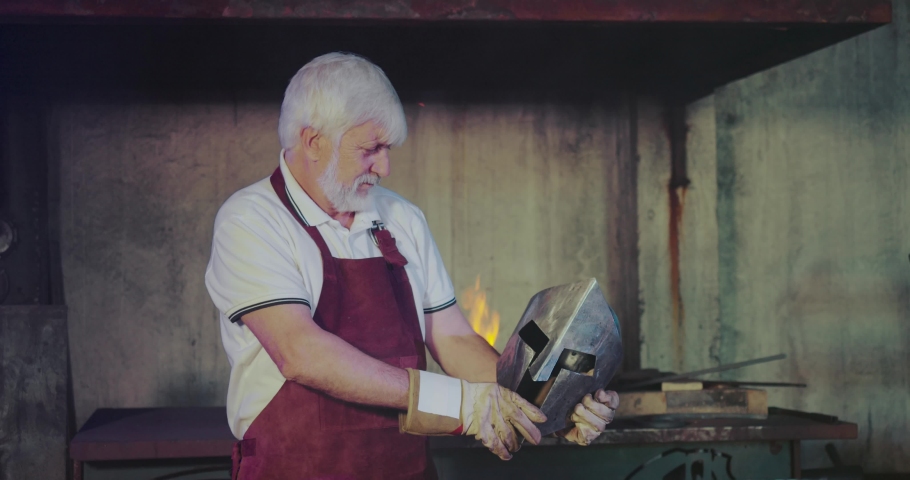 Front view of blacksmith steel black helmet holding. Male wearing red apron and gloves forging in smithy, looking, inspecting knight mask. Concept of craftsmanship and handwork.