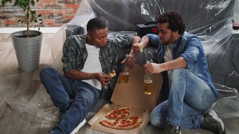 African-American man talks to friend while eats fresh pizza sitting on floor near foiled sofa. Young black men discuss repairing works in new house