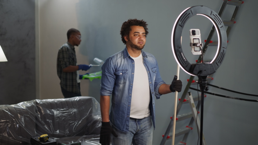 African-American man blogger shoots video while colleague applies paint on wall in studio. Black constructors teach audience to do domestic repair Royalty-Free Stock Footage #1090018801