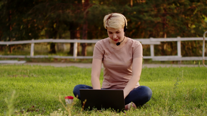 Woman with headset is sitting park on grass with laptop and says something into microphone and types on laptop, front view. Woman remote work in support service Royalty-Free Stock Footage #1090019957