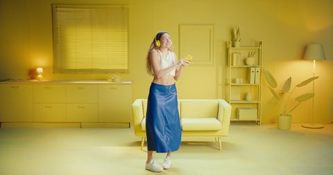 Slow motion of full length of cheerful young brunette woman girl in sleeveless top listens music with headphones from a mobile phone while dancing in the studio with monochrome yellow interior