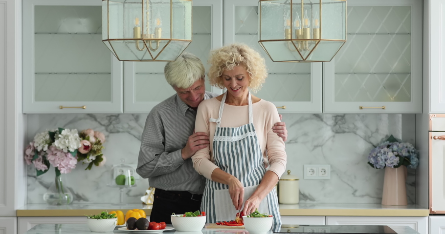 Loving aged wife prepare for older husband balanced vegetarian salad, hold knife cuts red paprika, standing together in kitchen talking enjoy cookery at modern home. Healthy lifestyle, cookery concept Royalty-Free Stock Footage #1090023323