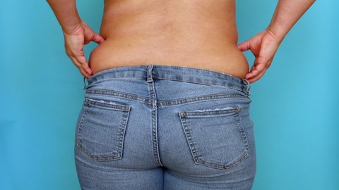 Back view of unrecognizable fat plump overweight woman wearing blue jeans, shaking, lifting, squeezing excess waist, massaging on blue background. Body positive, obesity, weight loss, liposuction.