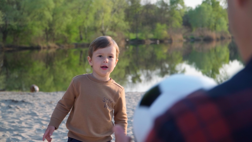 Little Boy Throws Soccer Ball to His Father. Man And Kid Playing With Black and White Football Ball Outdoors Near River. 4k