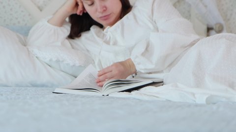 Young attractive woman in white retro-styled nightdress lies on the bed and reads a book in the bedroom at home, middle shot 4k video