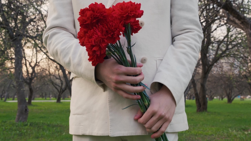 Young man in white suit outfit with buttons stands still and holds a bouquet of dark red carnations in his hands. Green grass in apple orchard in the city, sunset in spring. Slow motion dolly out.