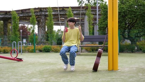 WROCLAW, POLAND - MAY 06, 2022: Lonely teenager kid boy drinking coca cola can sit swaying on a swing of playground