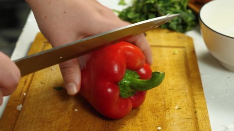 Close-up of a cook cutting red bell peppers on a kitchen board. In the background is a bunch of parsley and a cup. The concept of delicious and healthy food
