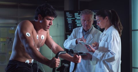 Cinematic shot of young athlete with medical electrodes doing endurance test of body resistance by training on gym cycle while physicians monitoring heart rate, pulse and ECG data in modern laboratory