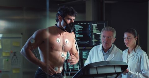 Cinematic shot of young athlete with medical electrodes doing endurance test of body resistance by training on treadmill while physicians monitoring heart rate, pulse and ECG data in modern laboratory