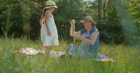 Slow motion of a happy little Caucasian girl playing bubble games with her pregnant mother outdoors in a park. Mother and daughter, blowing soap bubbles and enjoying summer holidays on the forest lawn