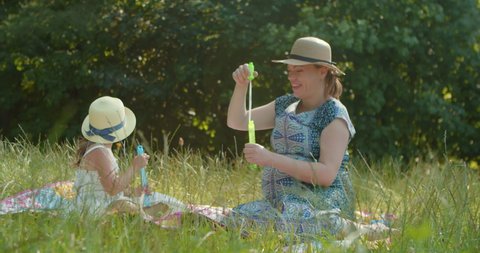 Pregnant mother and a young daughter are playing in a forest clearing on a hot summer day blowing soap bubbles from soap bubble sticks. Happy family spends their summer holidays in the park