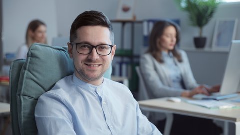 Close-up footage of male good-looking worker wearing black glasses sitting on comfortable designer chair in office. Portrait of handsome man in formal clothes looking at camera, smiling. Ordinary