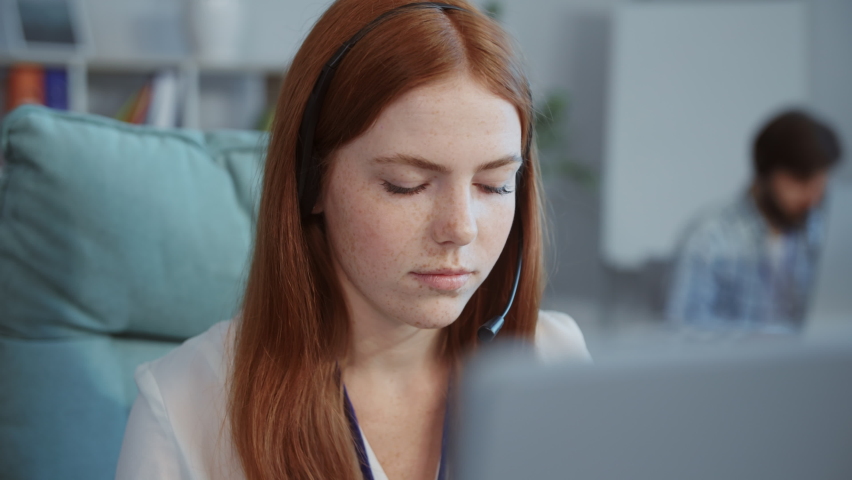 Close-up footage of redhead joyful woman with freckles sitting on chair raising hands to top. Happy Caucasian girl smiling. Positive emotions concept, good news. Blurred background with books. Office.