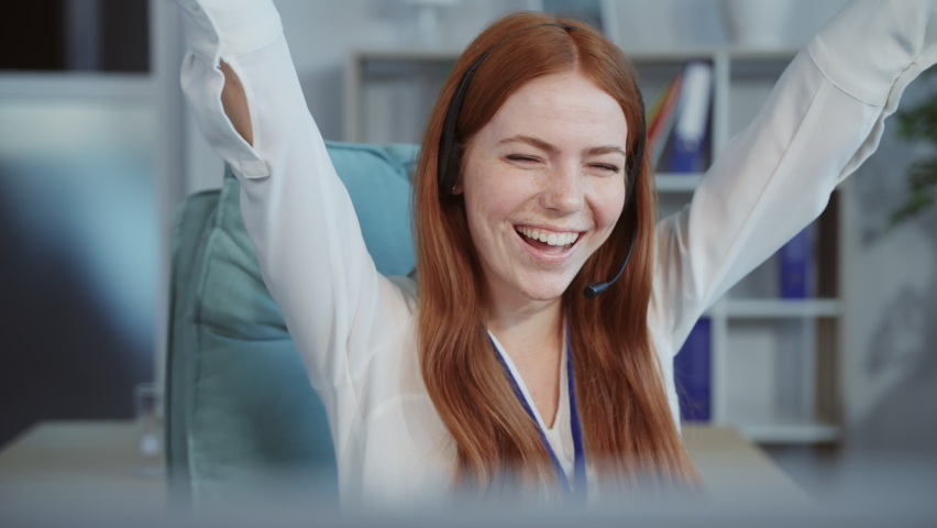 Close-up footage of redhead joyful woman with freckles sitting on chair raising hands to top. Happy Caucasian girl smiling. Positive emotions concept, good news. Blurred background with books. Office.