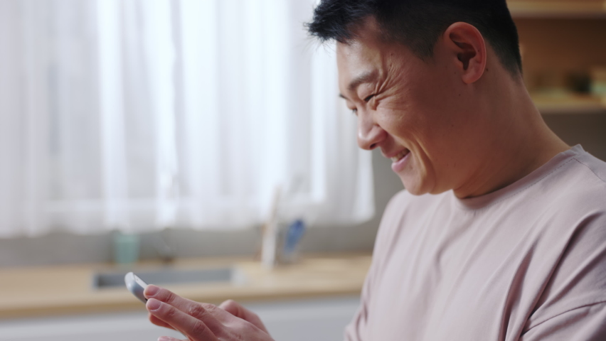 Close-up footage of smiling Asian man in casual clothes using mobile phone while cooking breakfast in cozy kitchen. Cheerful guy chatting with spouse. Online communication concept. Concept Royalty-Free Stock Footage #1090038005