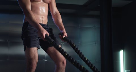 Cinematic shot of young muscular shirtless bodybuilder athlete is practicing endurance workout exercises for strength and concentration by training with lifting heavy battle ropes in gym.