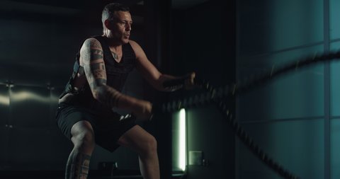 Cinematic shot of experienced muscular bodybuilder athlete with weight vest is practicing endurance workout exercises for strength and concentration by training with lifting heavy battle ropes in gym.